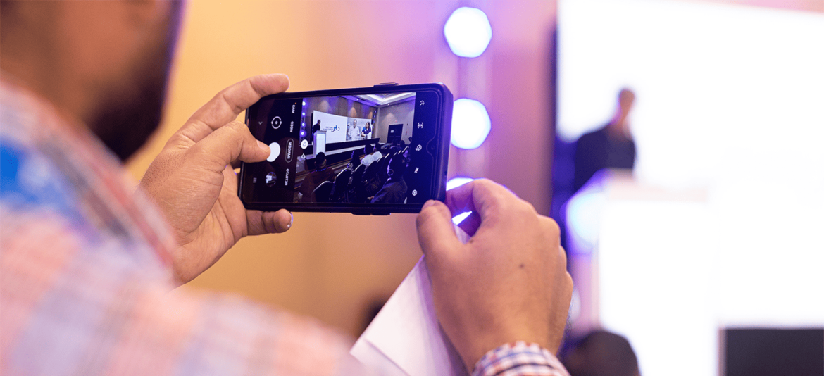 Un asistente sostiene un teléfono móvil, capturando una imagen de una presentación o conferencia en curso. En la pantalla del teléfono se observa una sala de conferencias con personas sentadas, mientras que en el fondo de la imagen real se ve al presentador desenfocado, de pie frente a un podio iluminado