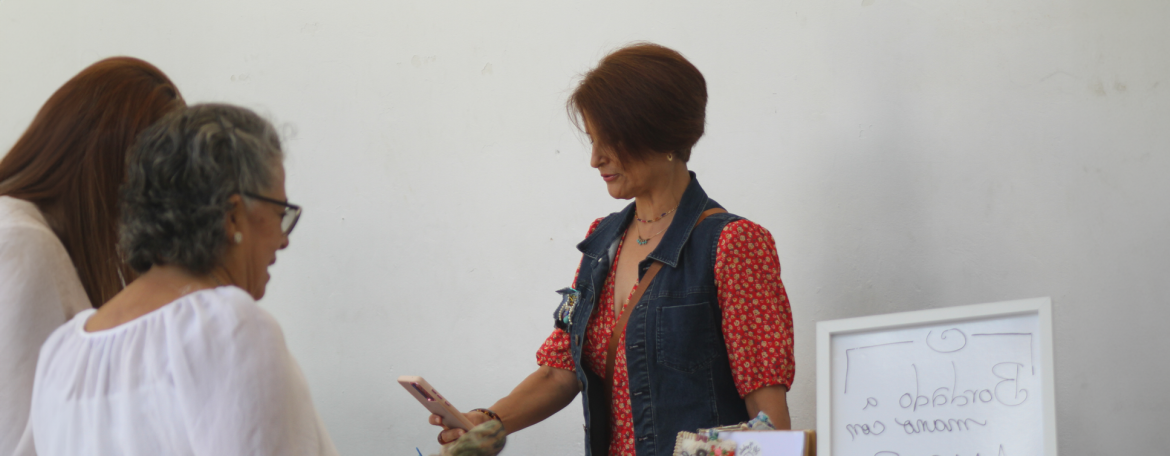 Una mujer con cabello corto y una blusa de flores rojas y chaleco de mezclilla está atendiendo a una cliente de cabello canoso y lentes. La mujer sostiene un teléfono móvil, aparentemente completando una transacción.