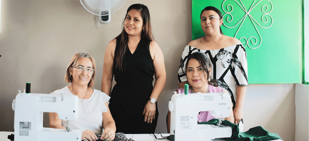 la-realidad-de-las-MYPES-lideradas-por-mujeres-en-l-salvador-entre-la-familia-y-el-negocio Cuatro mujeres están en un taller de costura, dos de ellas sentadas y usando máquinas de coser, mientras las otras dos están de pie detrás de ellas.
