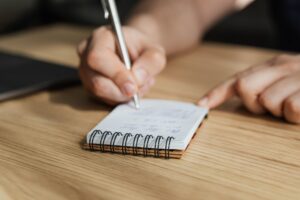 Mano de una persona escribiendo en una pequeña libreta de espiral sobre una mesa de madera, usando un bolígrafo