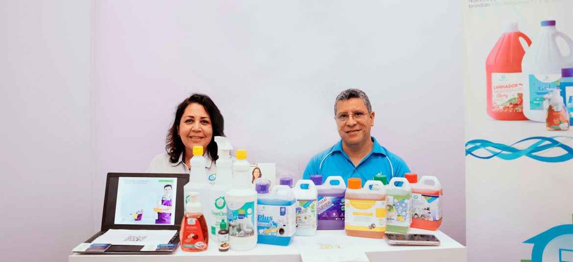 Dos personas, un hombre y una mujer, están de pie detrás de una mesa de exhibición con diversos productos de limpieza en botellas de diferentes tamaños y colores.