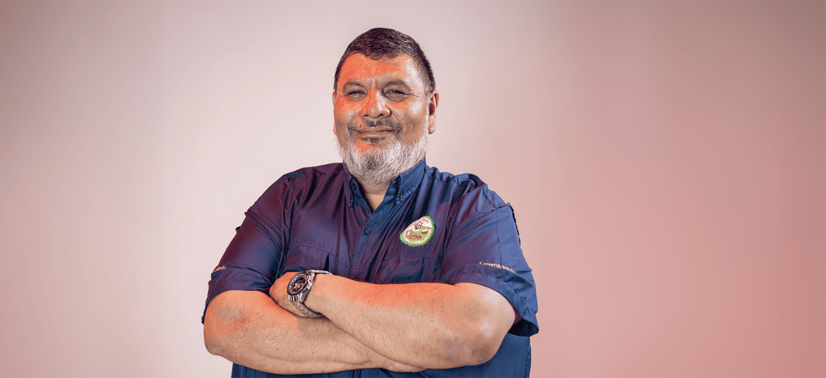La imagen muestra a un hombre de complexión robusta, con barba y cabello corto, cruzando los brazos frente a él. Está vestido con una camisa azul oscuro que lleva un logotipo bordado en el lado izquierdo del pecho, lo que podría representar su pertenencia a una empresa o negocio. Su expresión es de confianza y seguridad, lo cual refleja una actitud positiva y profesional.