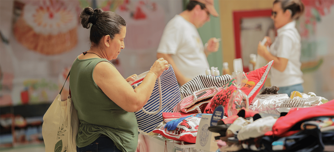 La imagen muestra a una mujer en un mercado o tienda observando detenidamente un producto, en lo que parece ser una selección cuidadosa de artículos. Lleva una bolsa de tela en el brazo y está rodeada de diversos productos exhibidos. Esta imagen refleja el proceso de toma de decisiones de un cliente, quien evalúa las opciones antes de realizar una compra.