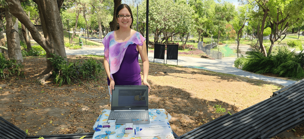 Una mujer de pie en un parque, sonriente y vestida con una blusa púrpura con un pañuelo de tonos pastel, está detrás de una mesa cubierta con un mantel de mariposas