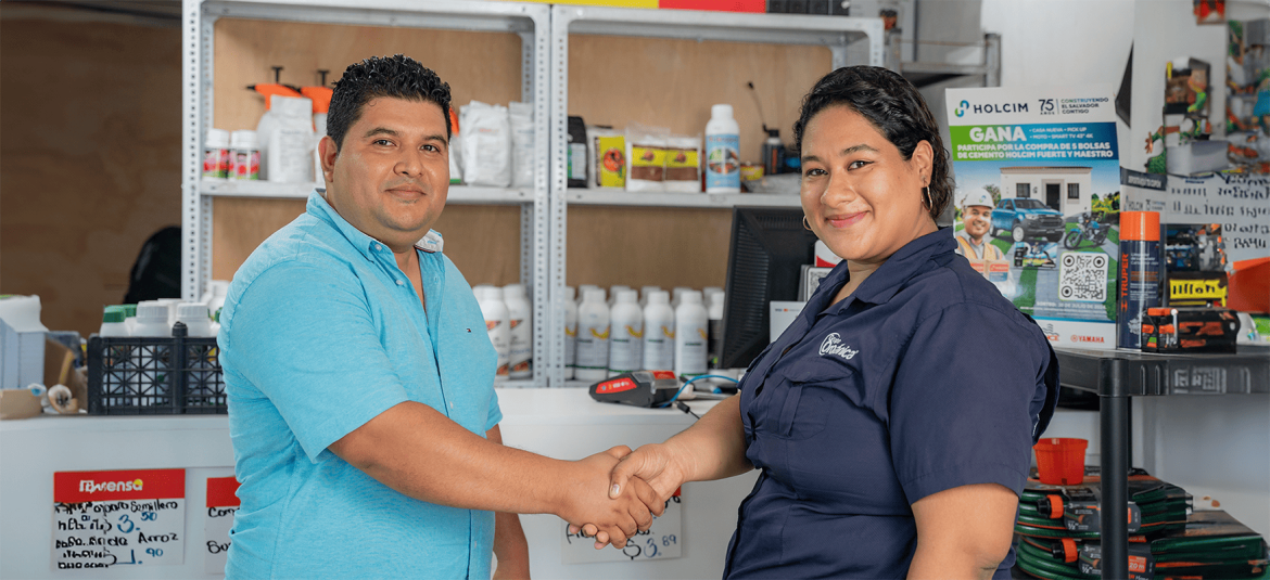 Un hombre y una mujer se dan la mano en un establecimiento comercial, sonriendo y proyectando una actitud amigable y de confianza. Él viste una camisa azul claro, y ella una camisa azul oscuro con el logo de su empresa. Detrás de ellos, se pueden ver productos en estantes y anuncios promocionales, sugiriendo un entorno de negocio o punto de venta. La imagen transmite un momento de colaboración, acuerdo o servicio al cliente, ideal para ilustrar temas de relaciones comerciales, alianzas o atención personalizada.