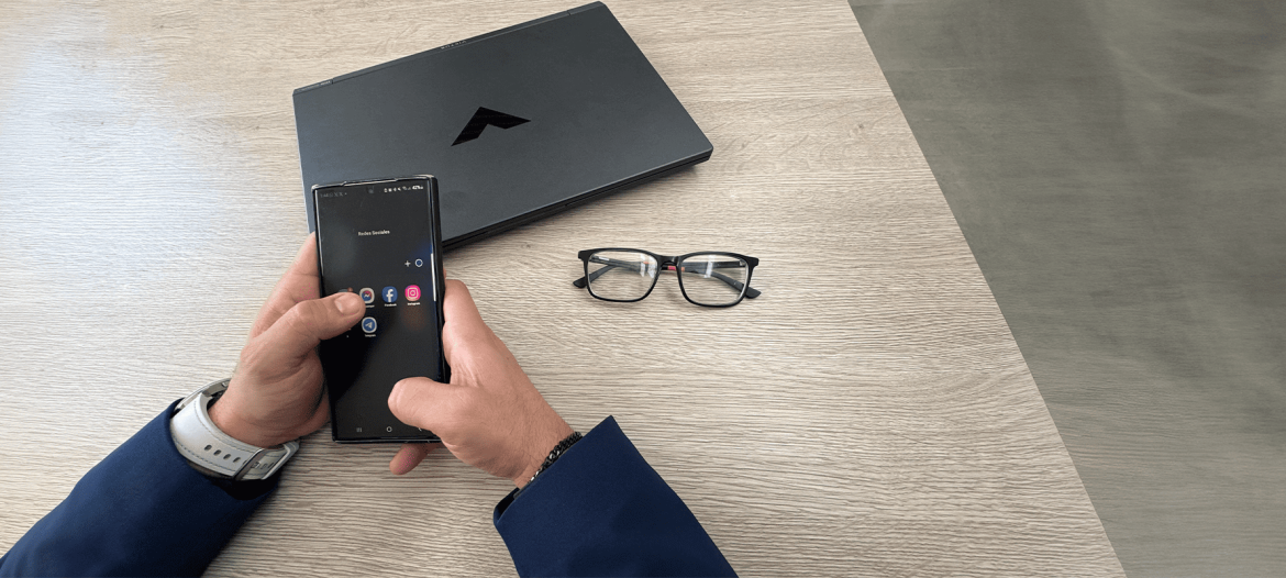redes-sociales-cual-es-la-más-conveniente-a-mi-negocio Un hombre con un reloj en la muñeca sostiene un teléfono móvil que muestra una pantalla con varias aplicaciones de redes sociales, como Facebook, Instagram y otras, mientras está sentado en un escritorio de madera clara. Sobre el escritorio hay unas gafas y una laptop cerrada con un logotipo oscuro en la tapa. La imagen transmite un ambiente de trabajo moderno y digital, enfocado en la tecnología y la conectividad a través de redes sociales.
