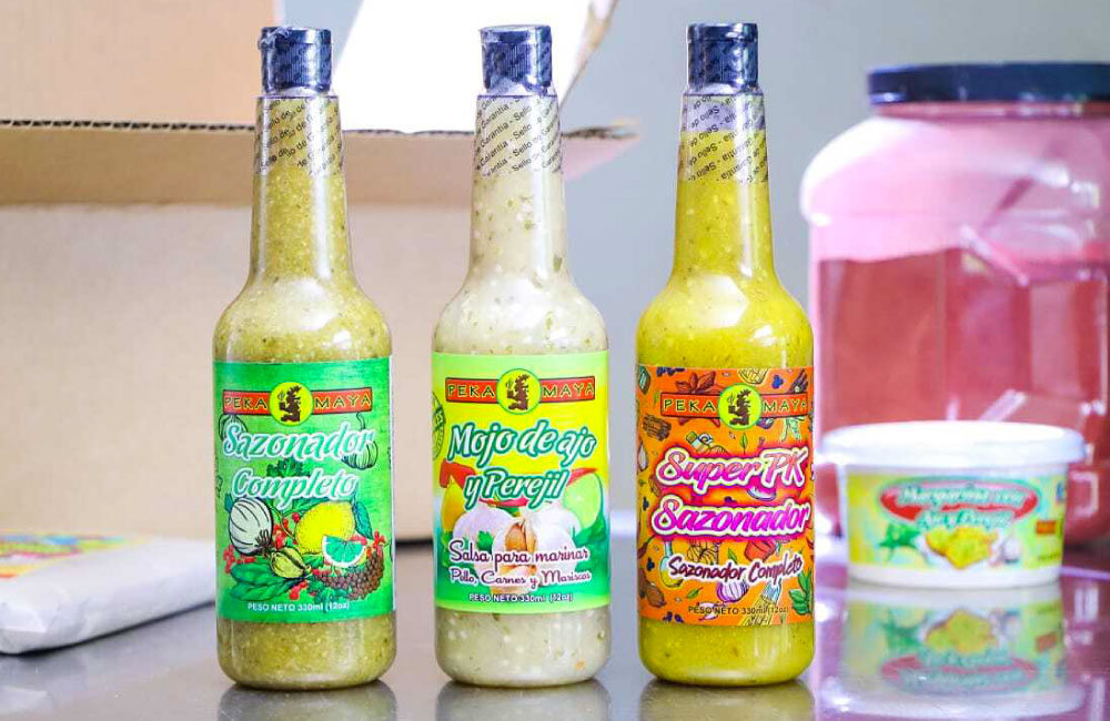 Botellas de salsas y sazonadores marca Peka Maya en una superficie, con etiquetas coloridas que indican los sabores 'Sazonador Completo', 'Mojo de ajo y perejil', y 'Super PK Sazonador'. En el fondo, se observan otros productos alimenticios y un envase grande.