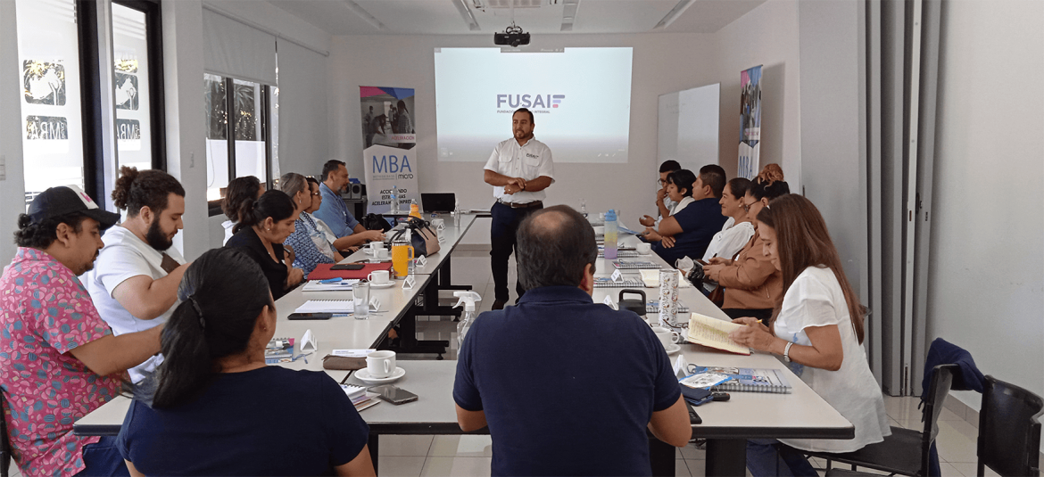 La imagen muestra a un grupo de personas en una sala de reuniones, participando en una sesión o taller organizado por FUSAI. Los participantes están sentados alrededor de una mesa rectangular, con materiales como cuadernos, tazas de café y botellas de agua frente a ellos. Al fondo, un facilitador o instructor está de pie, dirigiéndose al grupo, mientras una presentación con el logo de FUSAI se proyecta en la pantalla. Se observan carteles en las paredes con mensajes relacionados al MBA y al desarrollo empresarial, lo que sugiere que la sesión está enfocada en temas de capacitación o asesoría para emprendedores.