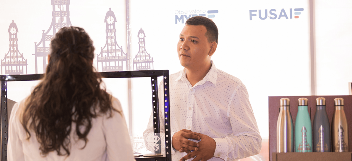 La imagen muestra a un hombre y una mujer en lo que parece ser una conversación dentro de un entorno profesional. Al fondo, se pueden ver los logotipos del "Observatorio MYPE" y "FUSAI". Además, hay botellas de agua reutilizables expuestas en una estantería, lo que sugiere un ambiente de feria o exposición de productos. La escena evoca una interacción relacionada con negocios o emprendimiento, posiblemente en un evento organizado para apoyar a las micro y pequeñas empresas.