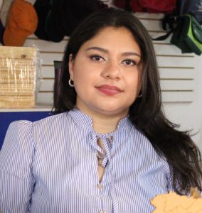 Retrato de una mujer joven de cabello largo y oscuro, vestida con una blusa de rayas azul claro con detalles en el cuello. Está de pie en un entorno de tienda o comercio, con estantes y productos al fondo. La mujer mira directamente a la cámara con una expresión calmada y amigable.