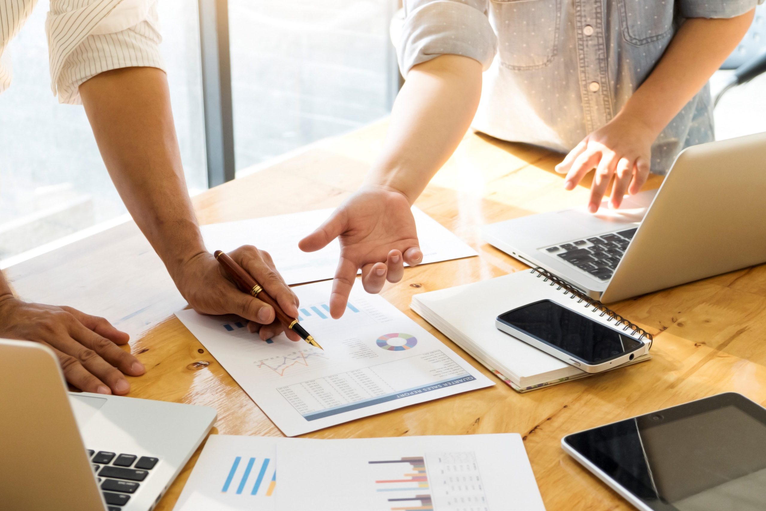 Personas colaboran en una mesa de trabajo, revisando gráficos y datos impresos. Una persona señala un gráfico con un bolígrafo, mientras otra gesticula, indicando una discusión o análisis.