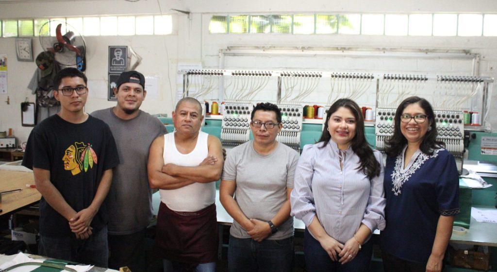 Fotografía de un grupo de seis personas en un taller. En la imagen, cuatro hombres y dos mujeres posan frente a máquinas industriales de bordado, con hilos de varios colores al fondo. Todos están de pie y miran a la cámara, algunos con los brazos cruzados y otros con las manos relajadas. La escena muestra un ambiente de trabajo colaborativo en una empresa de manufactura o bordado.