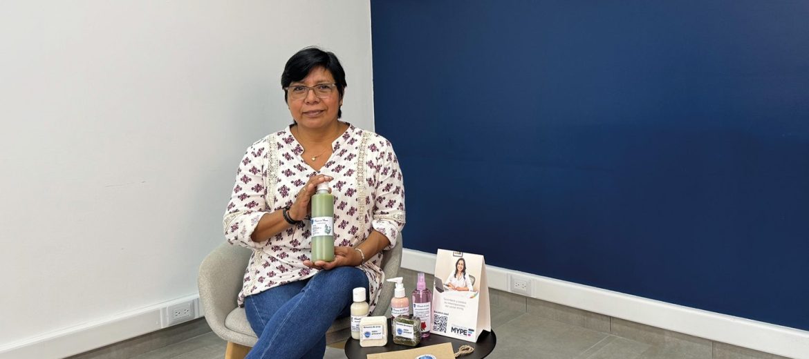 La imagen muestra a una mujer sentada en una silla, sosteniendo un producto en sus manos. Ella viste una blusa de estampado floral en tonos morados y lleva lentes. Frente a ella, sobre una mesa pequeña, se encuentran otros productos naturales, como cremas y lociones, junto a un cartel promocional que sugiere su participación en algún programa de apoyo para microempresarios. La escena transmite profesionalismo y dedicación, y parece que la mujer es una microempresaria enfocada en productos naturales, posiblemente elaborados de manera artesanal. Esta imagen refleja el esfuerzo de una emprendedora que, con el apoyo de iniciativas para MYPEs, impulsa su negocio en el sector de productos naturales.