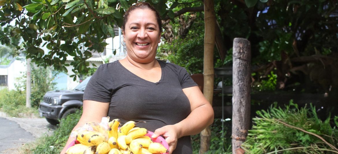 La imagen muestra a una mujer sonriendo, sosteniendo un recipiente lleno de bananos. La escena parece capturada al aire libre, en un entorno natural o rural, rodeado de vegetación y árboles. La expresión de la mujer refleja orgullo y satisfacción, posiblemente vinculados a su labor como microempresaria o productora agrícola. Esta imagen transmite una sensación de emprendimiento y autosuficiencia, destacando el trabajo de los microempresarios en áreas rurales o turísticas, quienes logran desarrollar sus negocios en armonía con su entorno. Podría estar relacionada con iniciativas de apoyo a pequeños productores, en las que se resalta la importancia de su trabajo para la economía local.