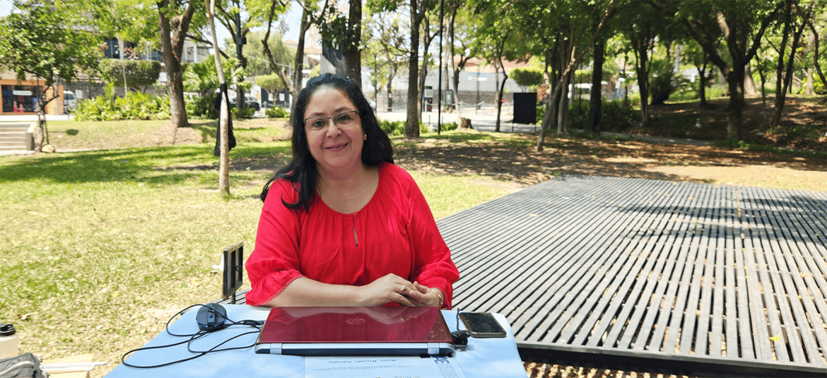la-tecnología-como-clave-del-exito-de-las-MYPEs La imagen muestra a una mujer sentada al aire libre, en lo que parece ser un parque o área verde. Ella está frente a una mesa que tiene sobre ella una laptop, un teléfono y algunos otros objetos. Lleva una blusa roja y tiene una expresión relajada y amable. Al fondo se observan árboles, arbustos y un espacio abierto, lo que aporta un ambiente natural y tranquilo a la escena. La luz natural sugiere que la foto fue tomada durante el día. La imagen podría representar a una profesional o empresaria trabajando en un ambiente externo, disfrutando de un espacio al aire libre.