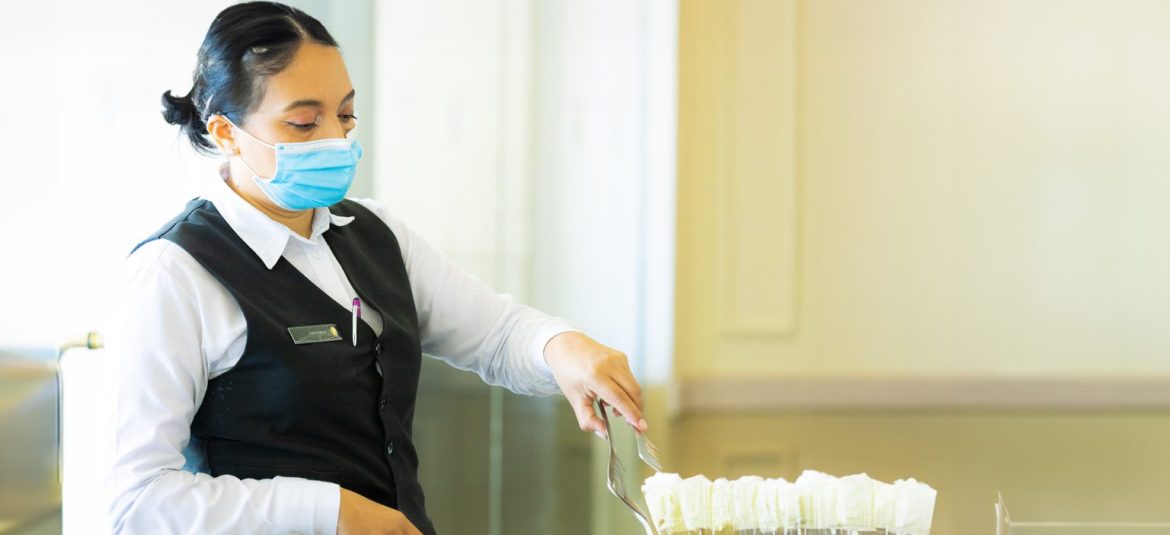mujer-sirviendo-comida La imagen muestra a una empleada de servicio vestida con uniforme, usando mascarilla facial y sirviendo comida con unas pinzas. La trabajadora parece estar en un entorno de restaurante o bufé, siguiendo protocolos de seguridad e higiene.