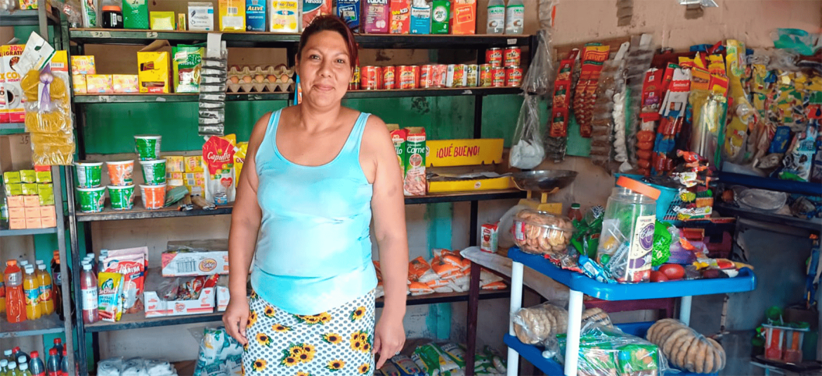 La imagen muestra a una mujer dentro de una tienda o pequeño negocio de abarrotes. Ella está de pie frente a un estante que contiene varios productos de consumo básico, como alimentos enlatados, bebidas, y paquetes de alimentos secos. La mujer lleva una blusa sin mangas de color celeste y una falda con estampado de girasoles. Su expresión es relajada y amigable. Al fondo, los estantes están organizados con diferentes artículos de uso diario, como paquetes de cereales, sopas instantáneas, galletas y otros productos. En la esquina inferior derecha, hay un estante adicional que contiene bolsas de pan y otros alimentos empaquetados. La tienda parece sencilla y acogedora, representando un negocio típico de una microempresaria que ofrece productos básicos a la comunidad local.