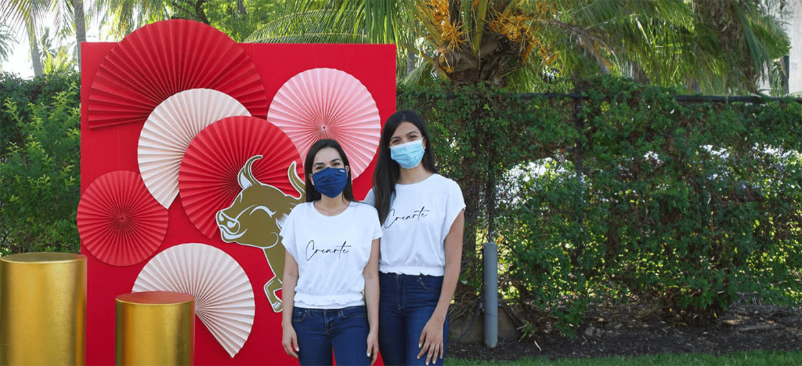 La imagen muestra a dos mujeres de pie al aire libre, ambas usando mascarillas y camisetas blancas con el texto "Crearte". Están frente a una decoración de fondo rojo adornada con abanicos de papel en tonos rojo y blanco, y un diseño de toro en el centro. También hay columnas doradas decorativas en la escena. El ambiente es luminoso, y detrás de ellas se pueden ver árboles y vegetación, lo que sugiere un evento o celebración al aire libre. La decoración y la vestimenta de las mujeres crean un ambiente alegre y organizado, posiblemente relacionado con un proyecto artístico o cultural.