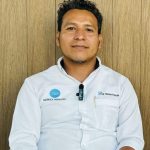 Hombre salvadoreño sentado frente a una pared de madera, vestido con camisa blanca con los logotipos de Konica Minolta y Sistemas de Inspección, portando un micrófono de solapa. Mira a la cámara con expresión serena.