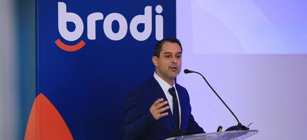 Un gerente en traje azul dando un discurso en un evento de Brodi, con un fondo azul que muestra el logotipo de la marca Brodi. Se encuentra frente a un micrófono y un podio transparente.
