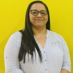 Mujer salvadoreña de cabello largo y lacio, con gafas y blusa a rayas azul claro, sonríe levemente frente a un fondo amarillo vibrante.