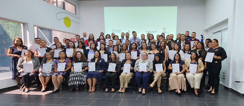 Grupo de 80 empresarios y empresarias celebrando su graduación en un evento organizado por la Escuela LID de FUSAI. Todos los graduados están sosteniendo sus diplomas y posando para la foto, con una bandera de El Salvador en el fondo y un ambiente de alegría y logro.