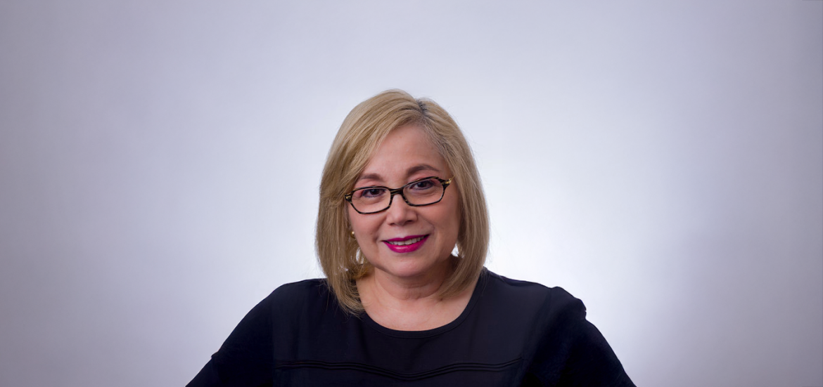 marketing-digital-en-la-mype-ana-maria-herrarte La imagen muestra a una mujer con cabello rubio, corto y liso, usando gafas y un vestido oscuro. Tiene un fondo de color liso, probablemente blanco o gris claro, y muestra una expresión serena con una ligera sonrisa.