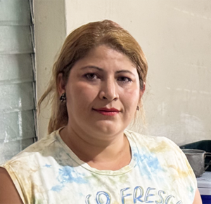 Fotografía de una mujer con cabello rubio y lacio, de tez clara, vistiendo una camiseta sin mangas con estampado en tonos pastel. Ella tiene una expresión seria y determinada, con labios pintados de rojo y aretes oscuros. Se encuentra en un espacio cerrado con paredes claras y una ventana con persianas metálicas en el fondo. La iluminación es artificial y resalta su rostro.