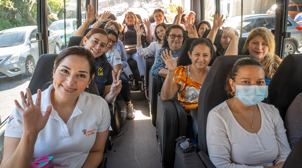 Un grupo de mujeres dentro de un autobús sonríe y saluda con la mano, reflejando un ambiente de entusiasmo y camaradería. Varias de ellas visten camisetas con logotipos de organizaciones como "Banco Integral" y "Escuela LID". Una mujer en primer plano usa mascarilla, mientras que otras muestran señales de alegría y optimismo. A través de las ventanas del autobús, se observa un entorno urbano con automóviles estacionados.