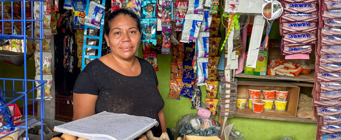 Fotografía de una mujer con cabello recogido y tez morena, vistiendo una blusa negra con detalles brillantes. Se encuentra en el interior de una tienda de abarrotes, rodeada de productos colgados y en estanterías, como golosinas, paquetes de fideos instantáneos, snacks y artículos de papelería. En primer plano, sostiene un cuaderno de cuentas abierto sobre un canasto con huevos. Su expresión es tranquila y segura. El fondo muestra las paredes verdes del negocio, con mercancía organizada en espacios reducidos.