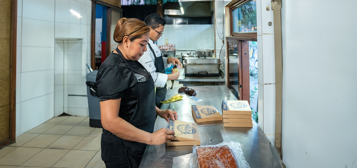 Dos trabajadores en una cocina industrial preparan y empaquetan productos horneados. La mujer en primer plano, vestida de negro, sella cajas de cartón con el logotipo de la marca, mientras que el hombre en el fondo, con uniforme de chef, manipula empaques dorados. En la mesa de trabajo hay varias cajas apiladas y un pastel envuelto en plástico.