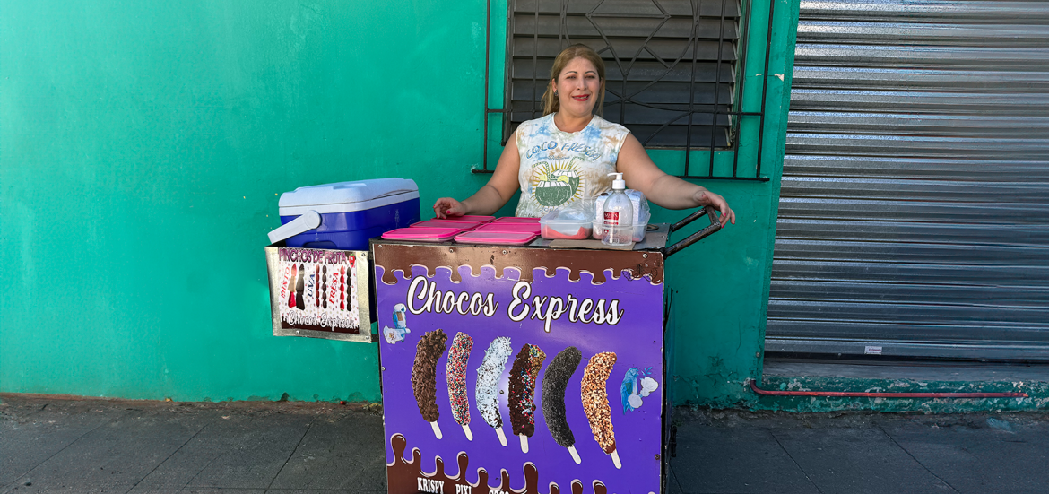 Mujer sonriente atiende su puesto de venta de "Chocos Express" en la calle, ubicado frente a una pared verde. Su carrito exhibe imágenes de bananos cubiertos de chocolate con distintos toppings. Sobre el mostrador hay recipientes plásticos, gel antibacterial y una hielera azul. La vendedora viste una camiseta sin mangas con un diseño tropical y muestra una actitud amigable y acogedora.