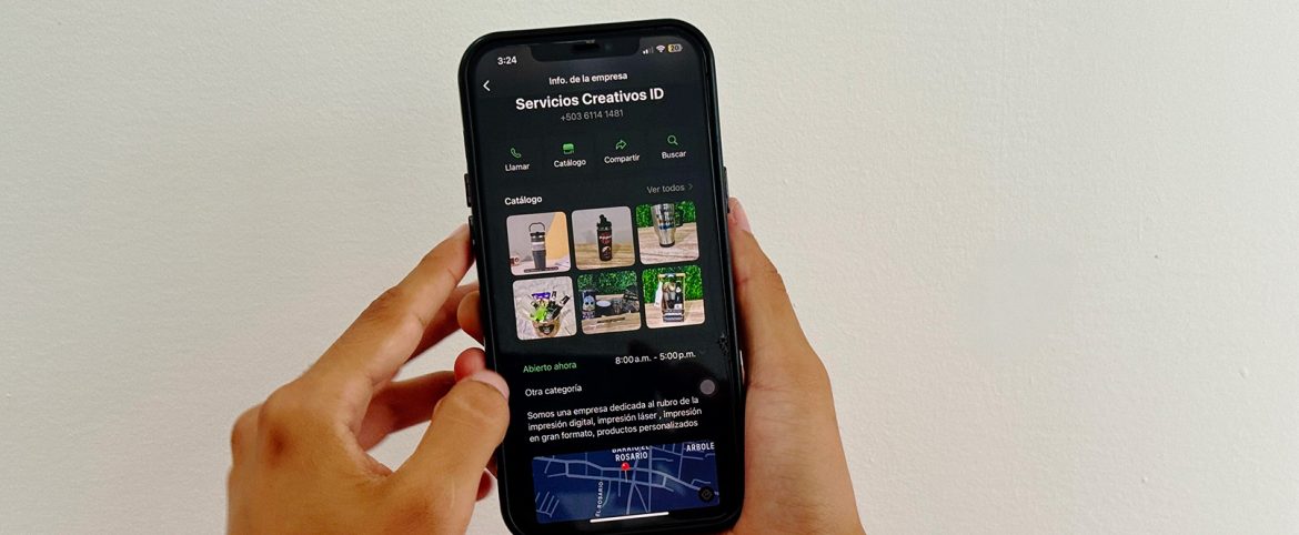 Una persona sostiene un teléfono inteligente con la pantalla encendida, mostrando la página de perfil de WhatsApp Business de 'Servicios Creativos ID'. En la pantalla se pueden ver opciones como 'Llamar', 'Catálogo', 'Compartir' y 'Buscar', junto con una galería de imágenes de productos personalizados, incluyendo botellas, vasos térmicos y kits de regalo. La empresa ofrece servicios de impresión digital y personalizada. Al fondo, una pared blanca proporciona un fondo limpio y minimalista.