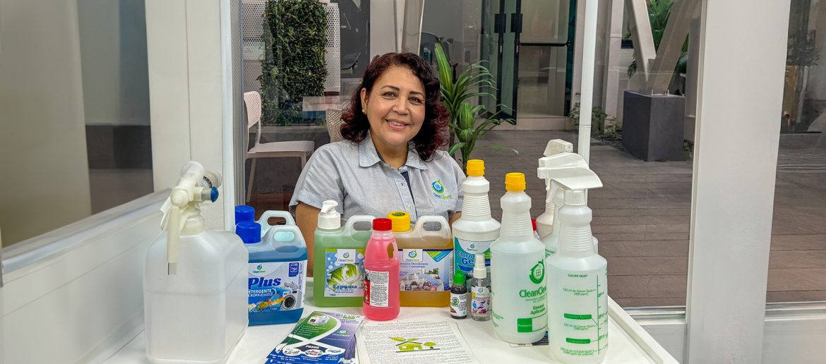Mujer de cabello rizado y camisa gris con logo empresarial sonríe detrás de una mesa con productos de limpieza. En la mesa hay botellas de detergente, desinfectante, cloro en gel y atomizadores con etiquetas de la marca 'CleanClever'. Detrás de ella, hay un espacio con ventanales que permiten ver un área con plantas y muebles de exterior.