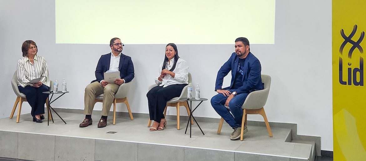 Cuatro panelistas participan en un foro sobre el sector MYPE en El Salvador. Están sentados en sillones grises en un escenario con una pantalla de fondo y mesas pequeñas con botellas de agua. La moderadora, una mujer de blusa a rayas, sostiene documentos mientras los otros panelistas escuchan. A la derecha, un hombre con chaqueta azul marino y jeans se inclina ligeramente hacia adelante. En el fondo, un cartel amarillo con el logo de la Escuela de Líderes MYPE (LID).