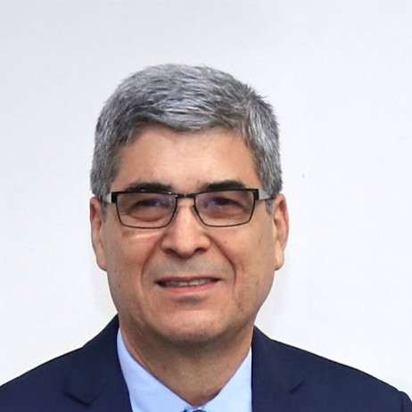 Retrato de un hombre de cabello canoso y corto, con gafas, vestido con saco azul marino, camisa celeste y corbata azul claro. Sonríe levemente mientras posa frente a un fondo blanco liso. La imagen tiene un estilo formal, tipo fotografía institucional o profesional.