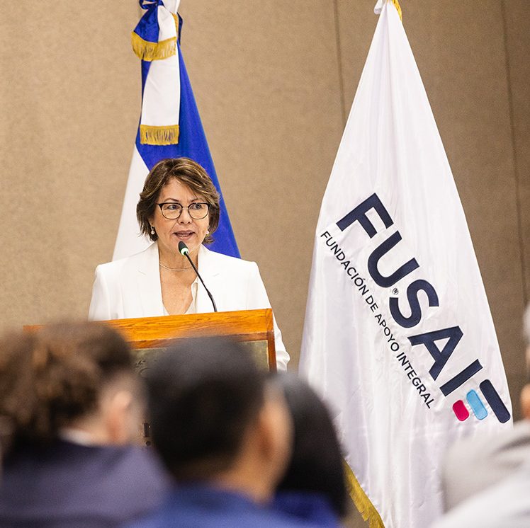 Mujer de mediana edad, con gafas y vestida con traje blanco, habla en un podio durante un evento institucional. A su lado se encuentran dos banderas: la de El Salvador y la de FUSAI (Fundación Salvadoreña de Apoyo Integral). En primer plano, se observa la audiencia de espaldas, escuchando con atención la intervención. El ambiente sugiere una conferencia o presentación formal.