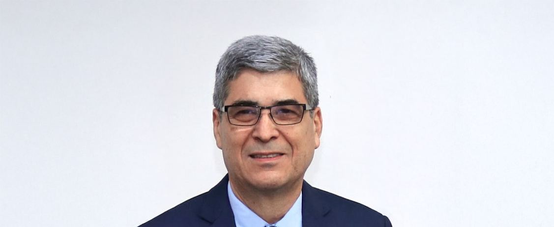 Retrato de un hombre de cabello canoso y corto, con gafas, vestido con saco azul marino, camisa celeste y corbata azul claro. Sonríe levemente mientras posa frente a un fondo blanco liso. La imagen tiene un estilo formal, tipo fotografía institucional o profesional.