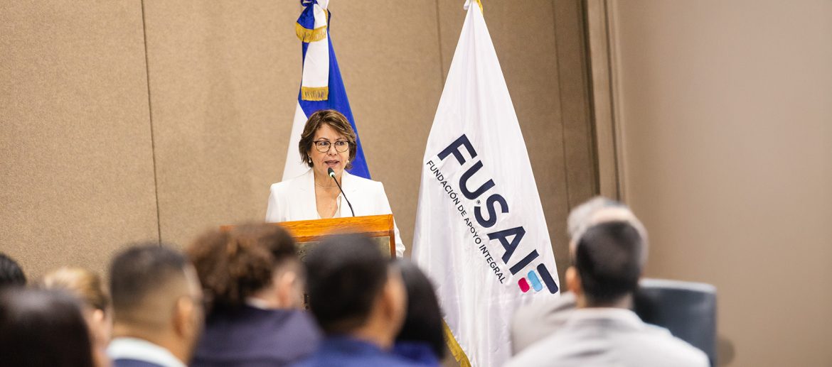 el-espejismo-del-emprendimiento-femenino-mas-negocios-menos-oportunidades Mujer de mediana edad, con gafas y vestida con traje blanco, habla en un podio durante un evento institucional. A su lado se encuentran dos banderas: la de El Salvador y la de FUSAI (Fundación Salvadoreña de Apoyo Integral). En primer plano, se observa la audiencia de espaldas, escuchando con atención la intervención. El ambiente sugiere una conferencia o presentación formal.