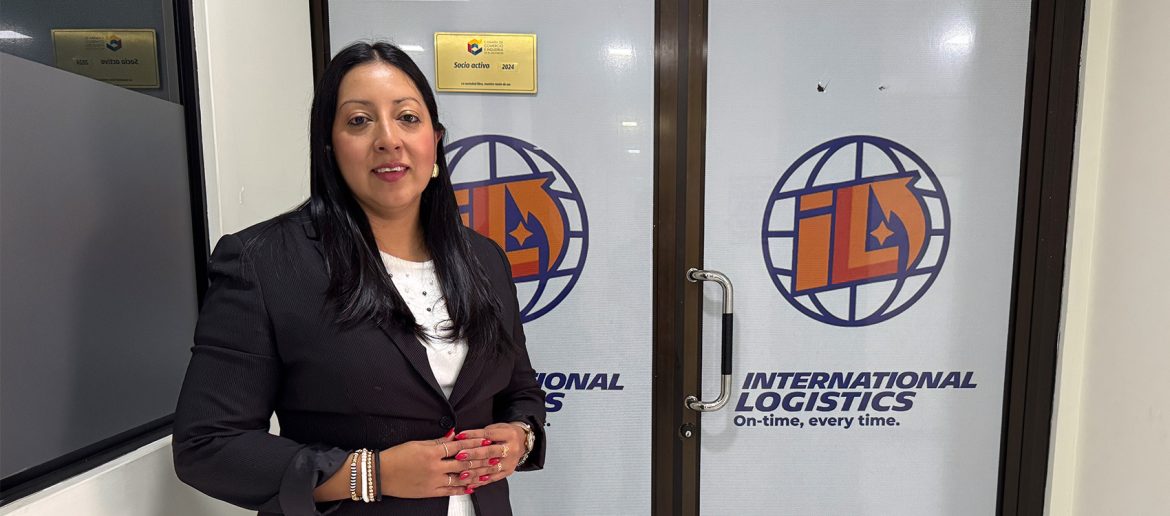 Mujer de cabello largo y lacio, vestida formalmente con saco oscuro y blusa blanca, posando frente a la entrada de una oficina. A sus espaldas se ve el logotipo de la empresa International Logistics, acompañado del eslogan "On-time, every time". En la puerta y en la pared contigua hay placas doradas que indican que la empresa es "Socio activo 2024" de la Cámara de Comercio e Industria de El Salvador. La mujer está sonriendo y con las manos entrelazadas al frente, mostrando una actitud profesional y segura.