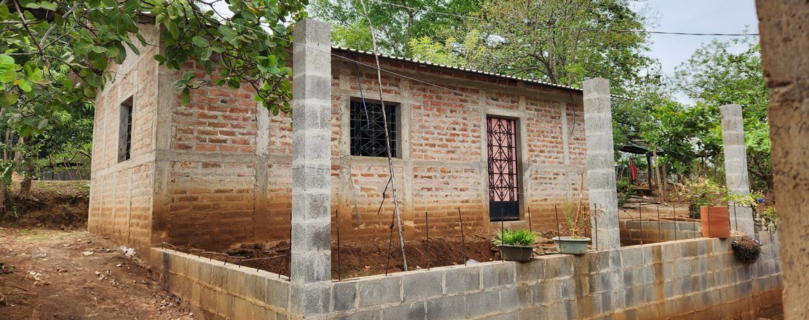 Vivienda de ladrillo en una zona rural rodeada de vegetación. La construcción es de una planta, con techo de lámina y ventanas con barrotes. Al frente, se observan columnas de concreto en proceso de construcción y varias macetas con plantas. El terreno tiene un ligero desnivel y está enmarcado por árboles y caminos de tierra. La imagen transmite un entorno en desarrollo con avances en infraestructura habitacional.