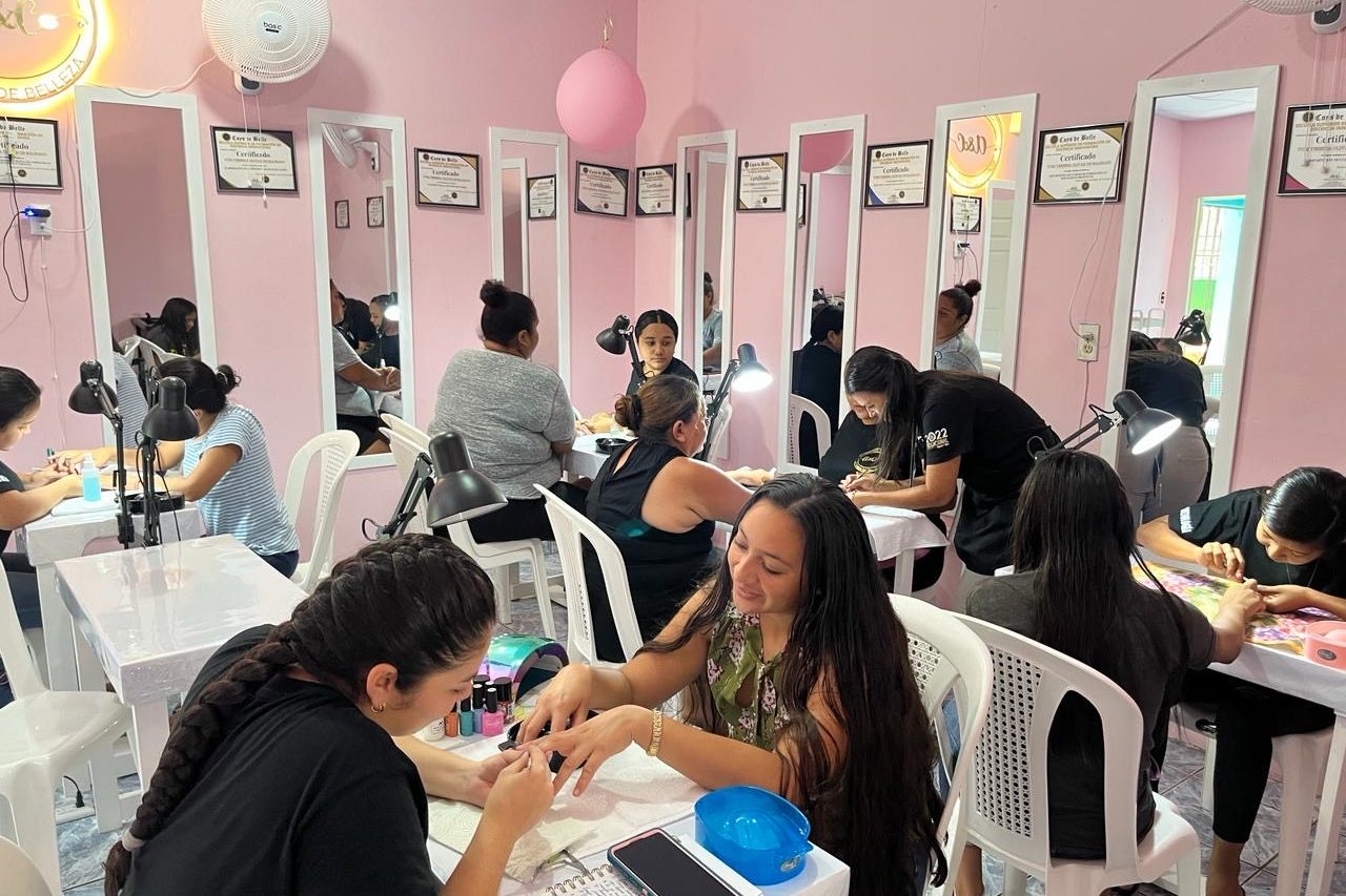 Salón de belleza con paredes rosadas donde varias mujeres están recibiendo servicios de manicura. Cada estación de trabajo cuenta con una lámpara y espejo individual, y en las paredes se observan múltiples certificados enmarcados. Las técnicas de uñas trabajan concentradas mientras las clientas conversan o sonríen, creando un ambiente activo, femenino y acogedor.
