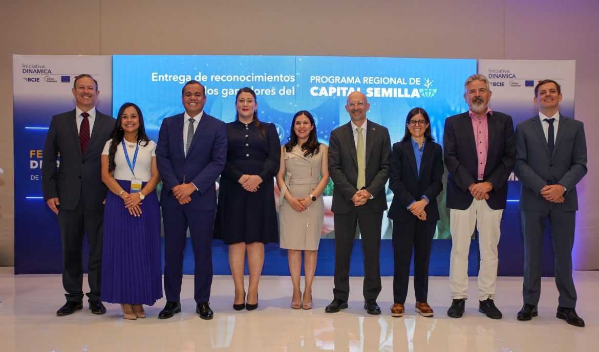 Grupo de nueve personas, hombres y mujeres, posando formalmente frente a un fondo institucional durante un evento de entrega de reconocimientos del “Programa Regional de Capital Semilla”, impulsado por la Iniciativa DINÁMICA del BCIE y la Unión Europea. El fondo azul incluye logotipos de las entidades organizadoras y el texto del evento. Todos los participantes visten ropa formal y sonríen a la cámara, en un ambiente profesional.