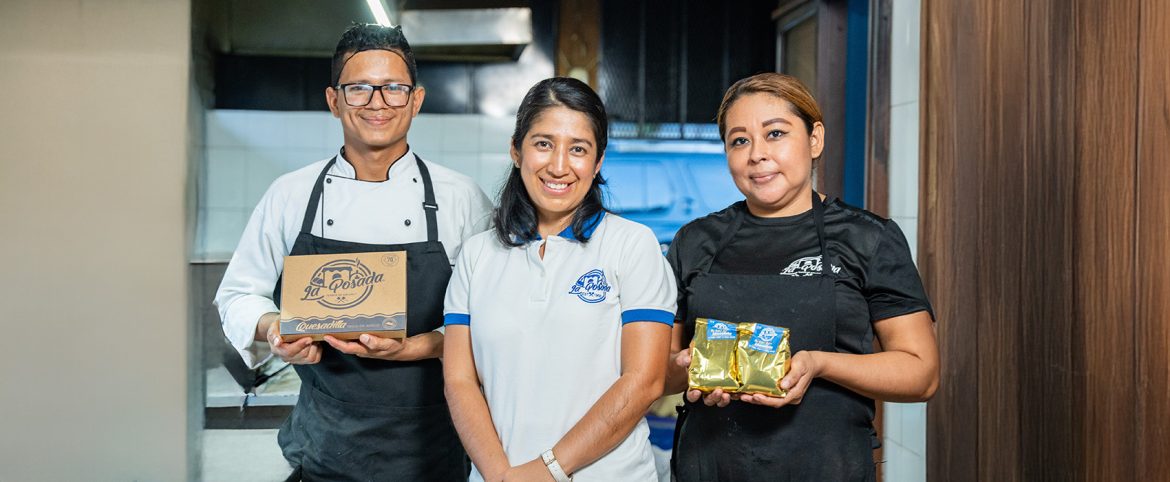 Tres personas posan en el interior de una cocina comercial. A la izquierda, un hombre con gafas, uniforme blanco y delantal negro sostiene una caja de productos con el logo "La Posada". En el centro, una mujer con cabello suelto y una camisa polo blanca con azul también lleva el logo "La Posada" bordado. A la derecha, otra mujer con delantal negro sostiene dos paquetes dorados, también etiquetados con el mismo logo. Todos sonríen y representan al equipo de un pequeño emprendimiento alimentario dedicado a la producción y venta de quesadillas.