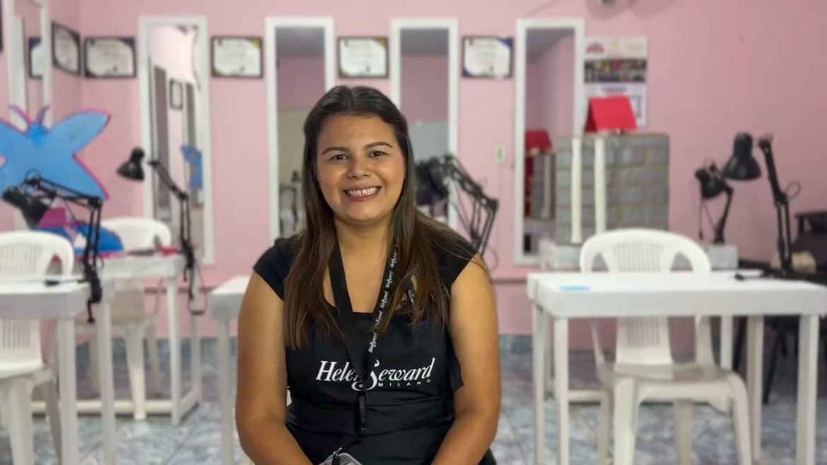 Mujer joven salvadoreña, sonriente, vestida con un delantal negro con el logo "Helen Seward Milano", sentada en un salón de belleza con paredes rosadas. Al fondo se observan espejos, lámparas y certificados colgados en la pared. La imagen transmite confianza y profesionalismo en un entorno dedicado a la estética.