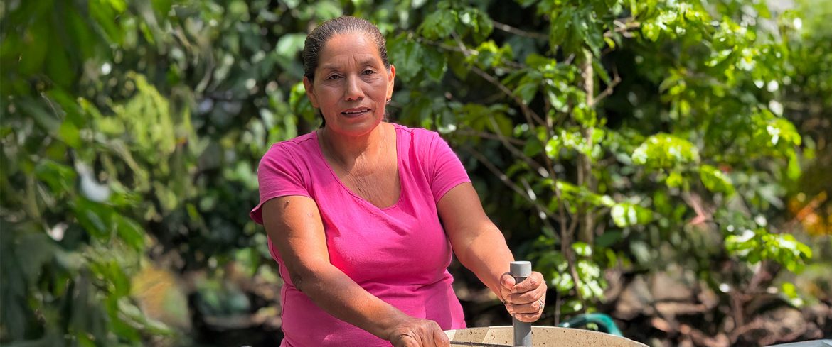 Mujer de mediana edad con camiseta rosa, trabajando al aire libre entre vegetación frondosa. Está utilizando una herramienta en un recipiente grande, posiblemente realizando una actividad relacionada con el reciclaje o la economía circular. La imagen transmite esfuerzo, conexión con la naturaleza y trabajo comunitario.