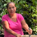 Mujer de mediana edad con camiseta rosa, trabajando al aire libre entre vegetación frondosa. Está utilizando una herramienta en un recipiente grande, posiblemente realizando una actividad relacionada con el reciclaje o la economía circular. La imagen transmite esfuerzo, conexión con la naturaleza y trabajo comunitario.