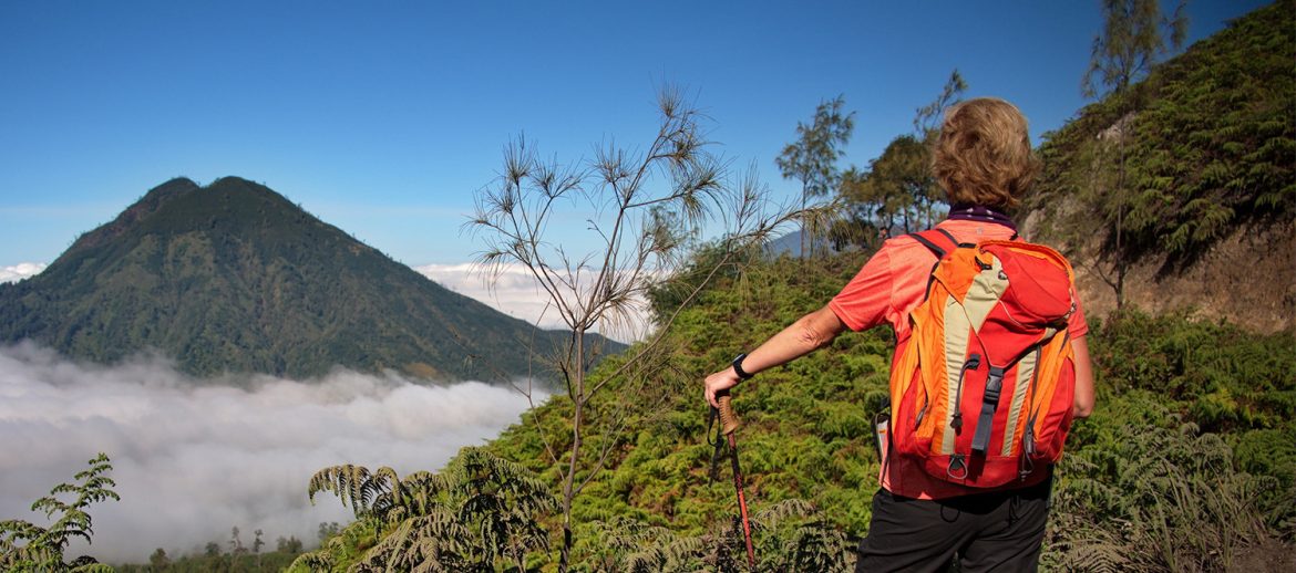 Persona de espaldas con mochila de senderismo naranja observa un paisaje montañoso. Frente a ella, un volcán cubierto de vegetación se eleva sobre un mar de nubes bajo un cielo azul despejado.