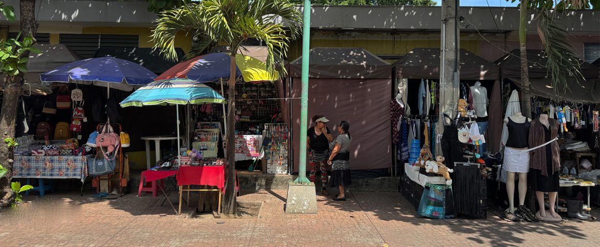 Puestos de venta informal ubicados en una acera urbana, con toldos de diferentes colores (azul, rojo, marrón y negro) que cubren mercadería variada. Se observan mochilas, peluches, bisutería, ropa colgada y maniquíes. Dos mujeres conversan frente a uno de los puestos, mientras otras personas no son visibles. Una palmera y un poste dividen visualmente la escena. La imagen muestra la actividad comercial de pequeños emprendedores en un entorno urbano y caluroso, con sombra de árboles.