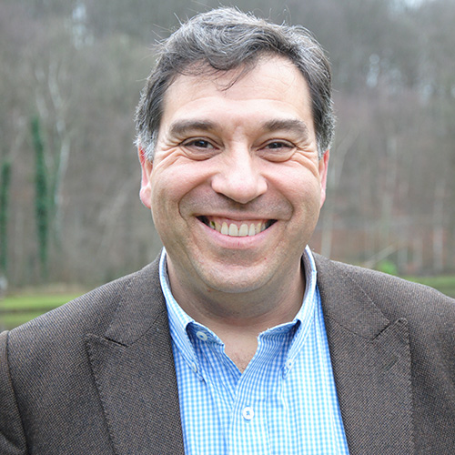 Retrato de un hombre de mediana edad, sonriente, de cabello corto y canoso, vistiendo saco marrón y camisa de cuadros azul claro. Al fondo se observa un paisaje natural con árboles sin follaje y tonos invernales desenfocados.